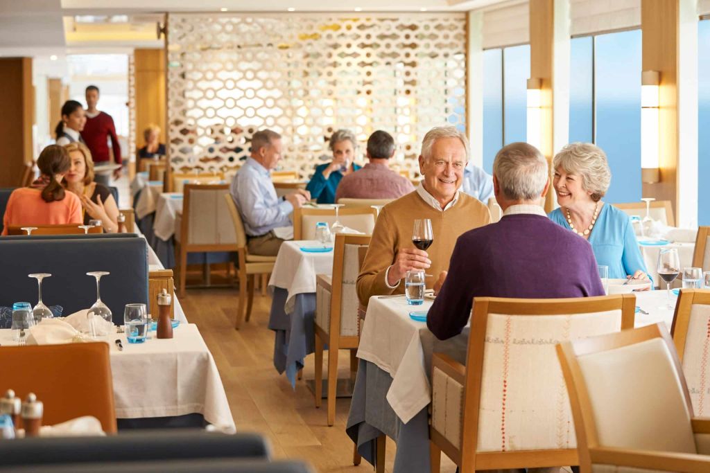 Ship restaurant with guests dining at white-clothed tables beside a lattice screen and sea-view windows.
