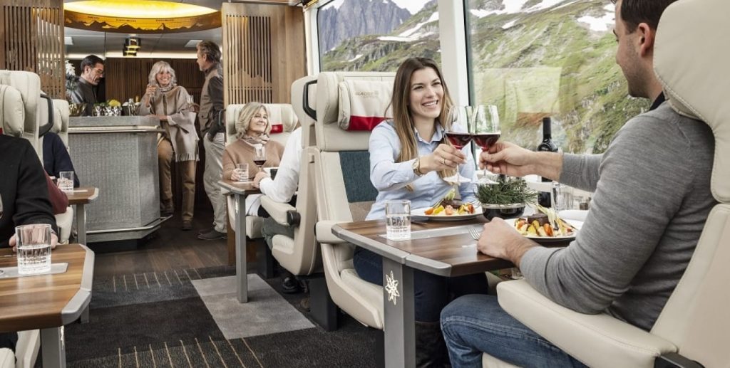 Diners sharing wine and plated meals at rail car tables beside a panoramic window with alpine peaks.