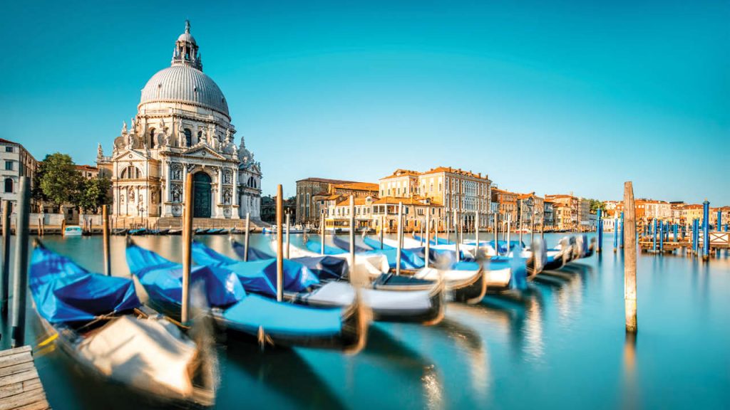 Domed basilica beside a canal lined with moored blue-covered gondolas.