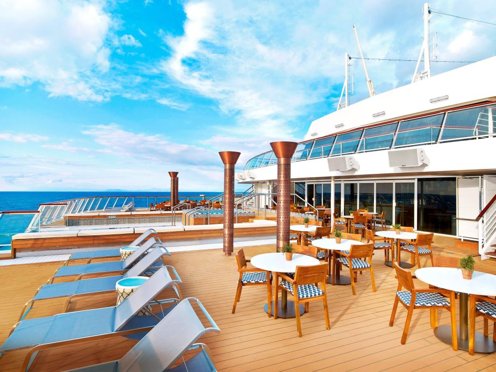 Ship deck with blue loungers, round wooden tables and copper columns beside a glass-walled bar.