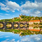 Stone arched bridge with statues across a river, with a hilltop fortress and reflections in calm water.
