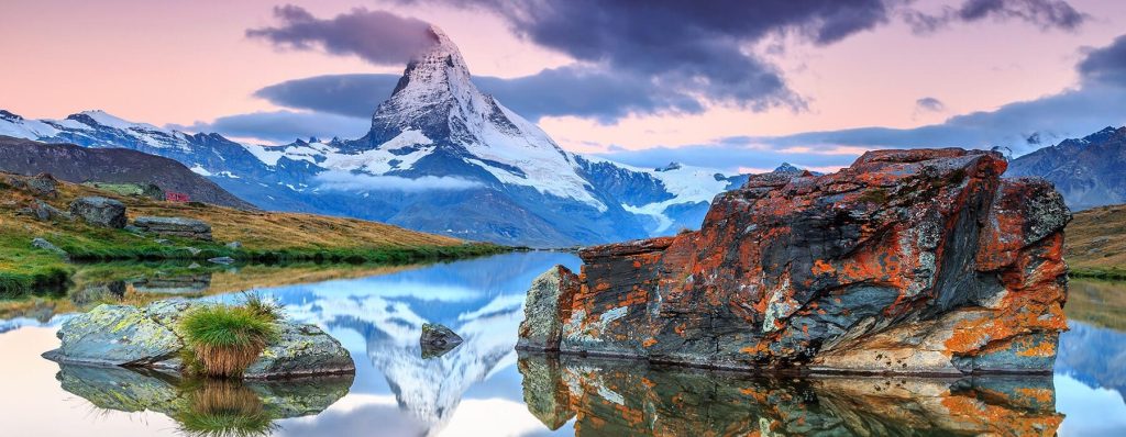 Matterhorn peak at dusk reflected in a still lake, with lichen-covered boulders in the foreground.