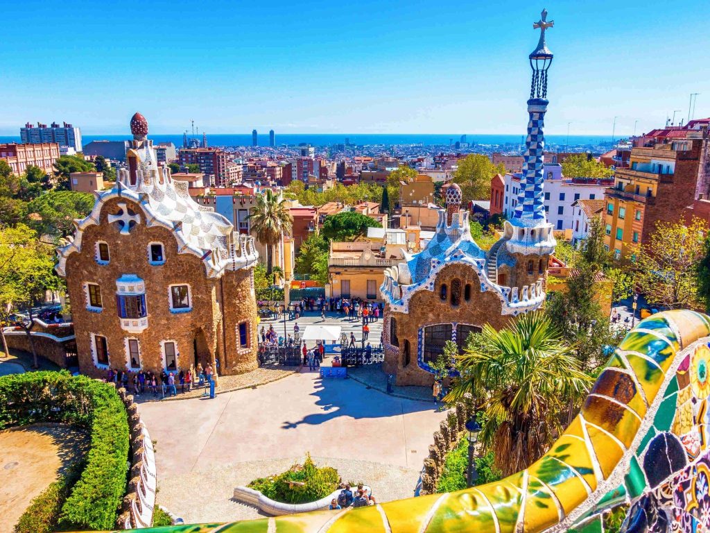 Ornate mosaic-tiled gatehouse pavilions with a palm tree and a hilltop view over a city and sea.