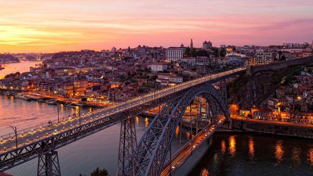 Illuminated arched steel bridge over a river at dusk, with a hillside old town and glowing reflections.