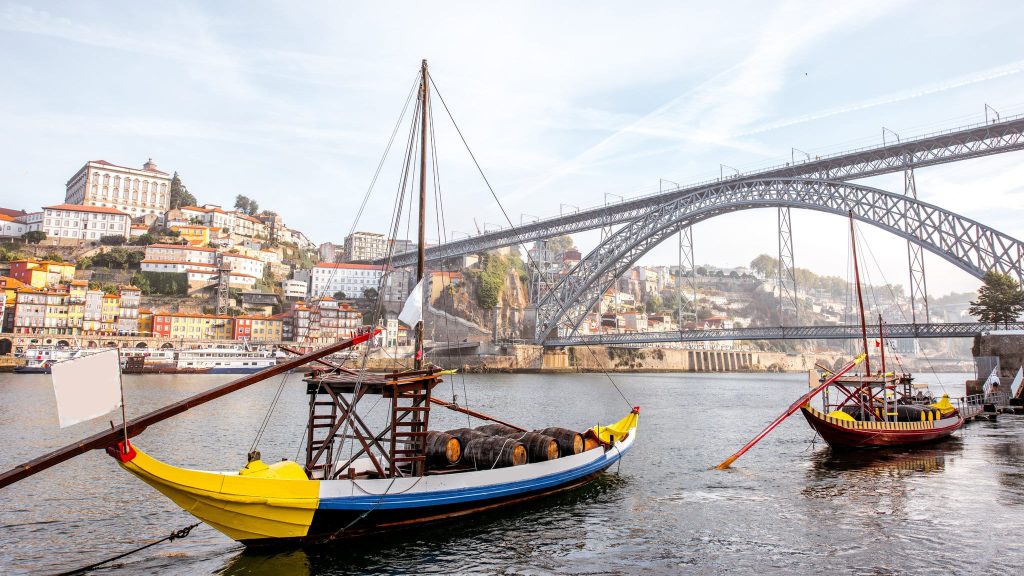 Traditional wooden boats with barrels and a sail moored on a river below a steel arch bridge.
