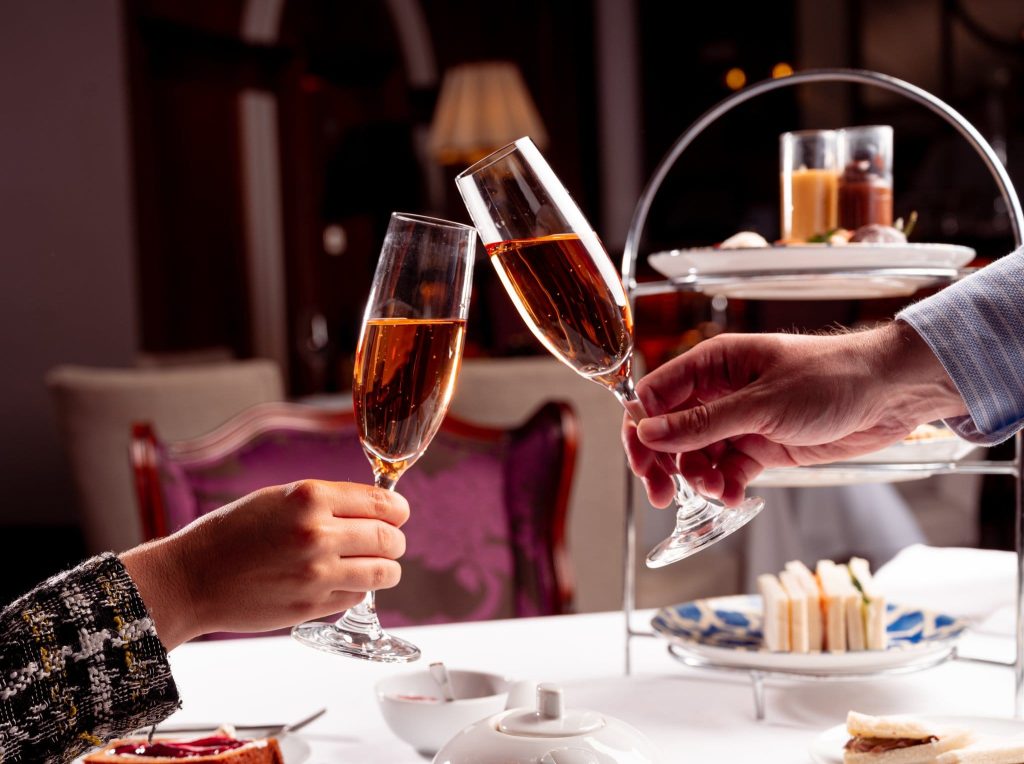 Champagne flutes being toasted at a table with an afternoon tea stand of sandwiches and pastries.