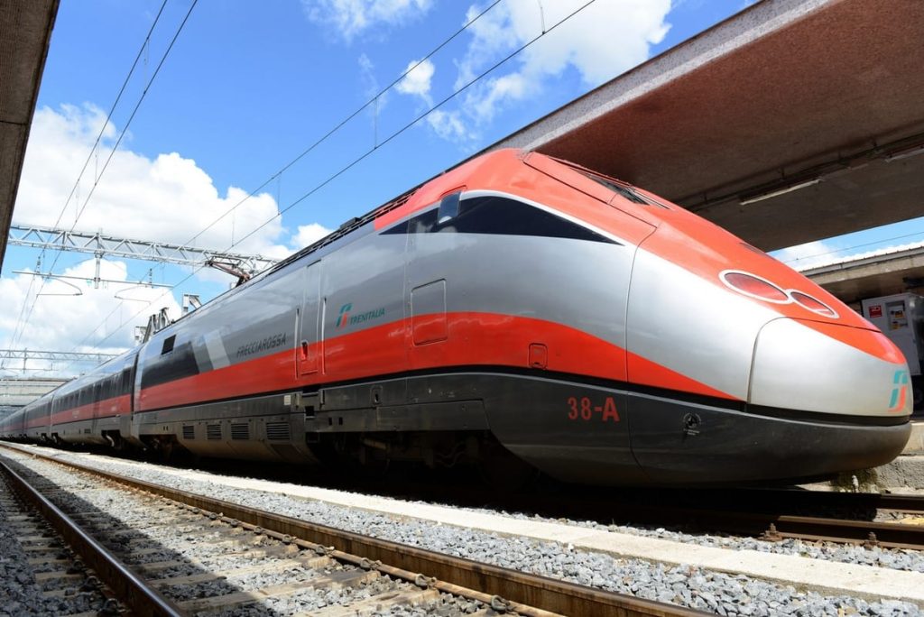 Red-and-silver high-speed Trenitalia train at a station platform under a blue sky.