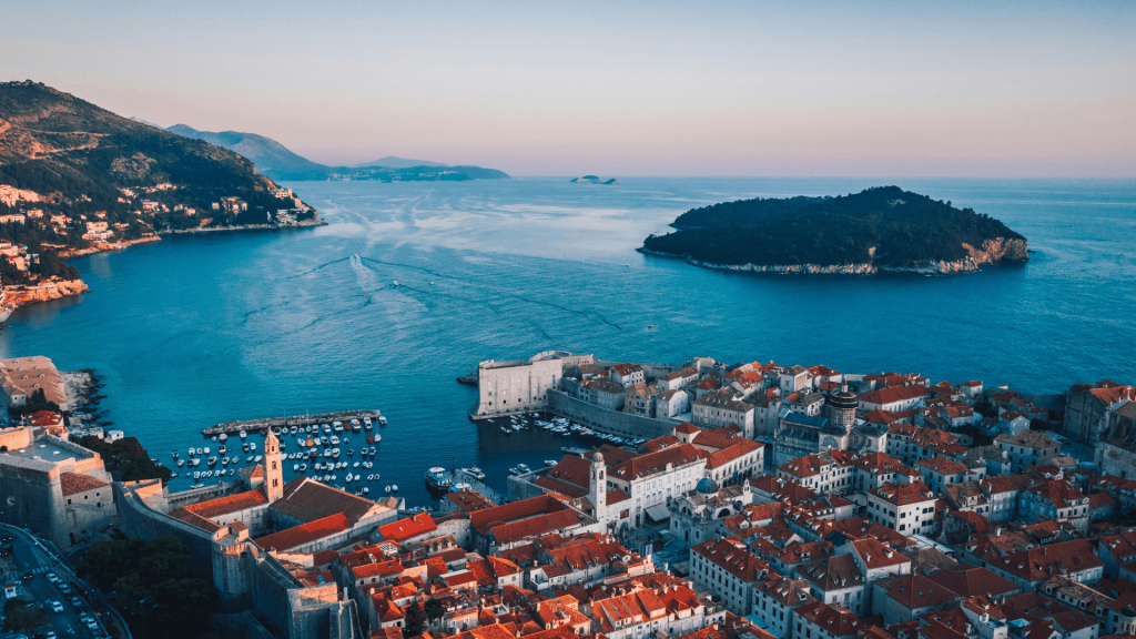 Aerial dusk view of a walled old town with red rooftops, a harbor, and an island in the sea.