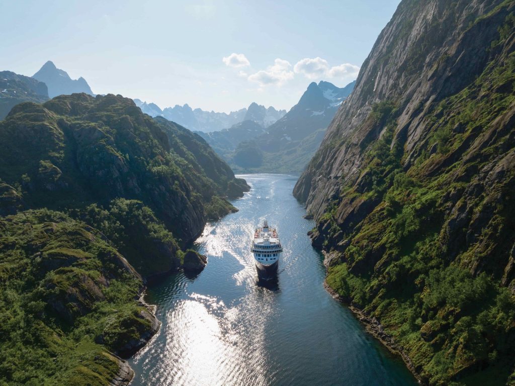 Small white expedition ship sailing between steep rocky cliffs in a narrow green fjord.