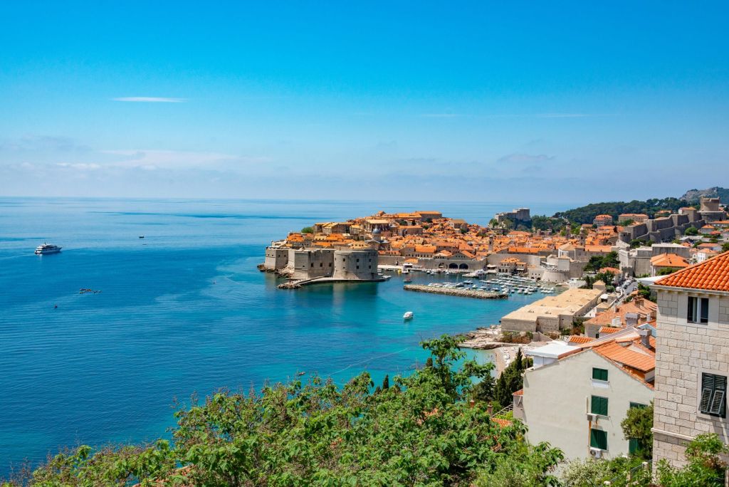 Walled old city with red-roofed buildings and stone fortresses on a sea bay under a clear blue sky.