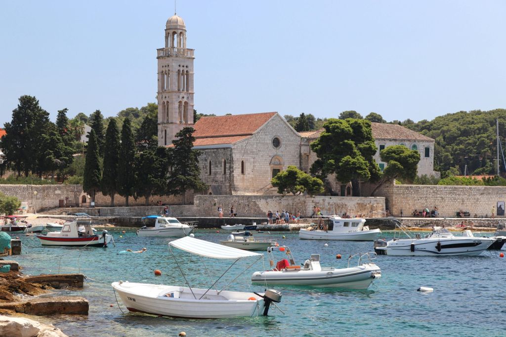 Small harbor with moored white boats in clear turquoise water and a stone bell-tower church behind.