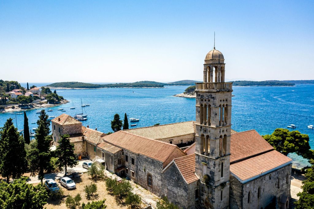 Stone monastery with a tall bell tower and terracotta roofs overlooking a bay with boats and islands.