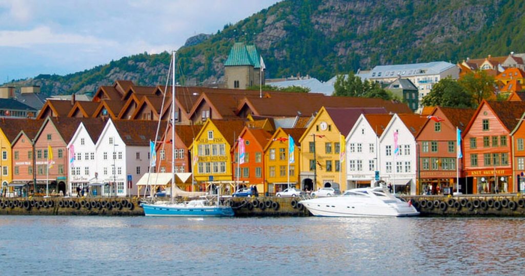 Colorful wooden waterfront buildings and moored boats on a harbor in front of a green hill.