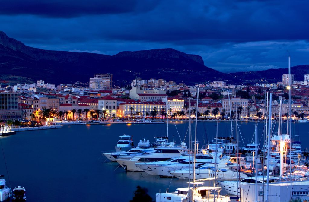 Dusk view of a harbor with luxury motor yachts moored in front of a hillside waterfront city.