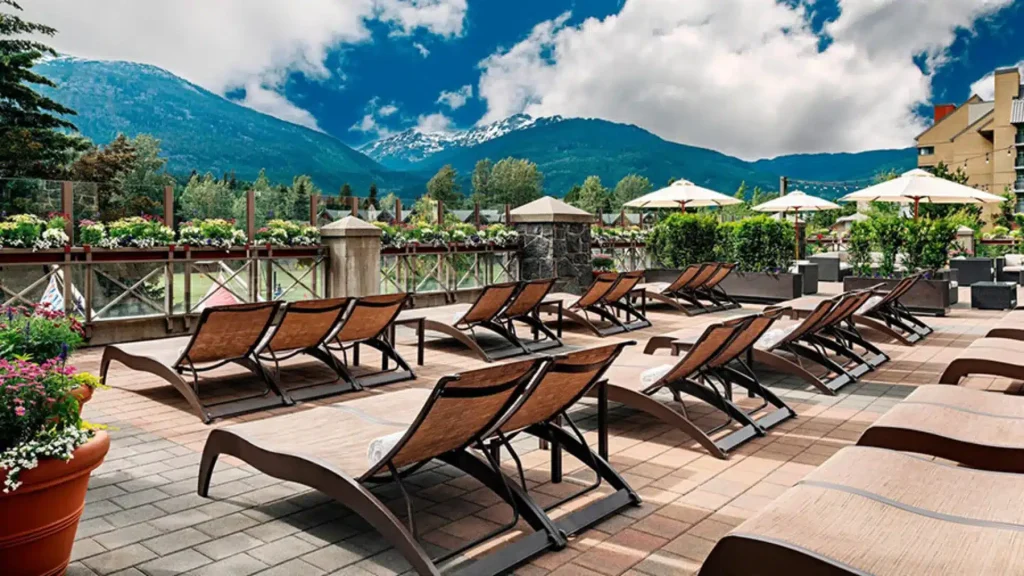 Hotel terrace with wooden lounge chairs, umbrellas, and potted flowers overlooking a mountain range in the distance.