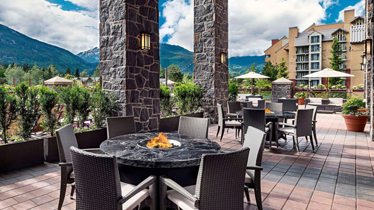 Outdoor hotel terrace with round fire pit tables, wicker chairs, and stone pillars, mountains in the distance.