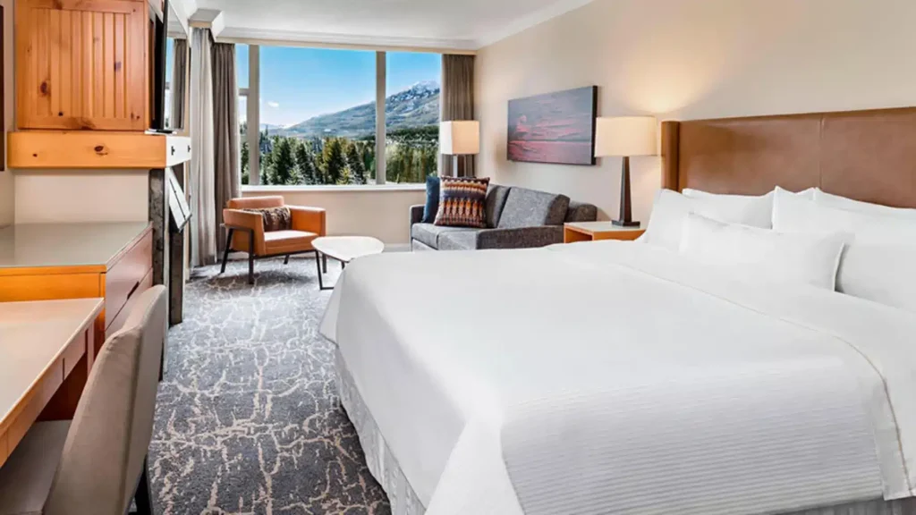 Hotel room with two white beds, a sofa and chairs by a large window showing snowcapped mountains outside.