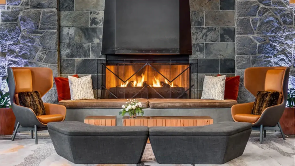 Cozy living room with a lit stone fireplace, four orange-brown lounge chairs, and a central coffee table with a flower arrangement.