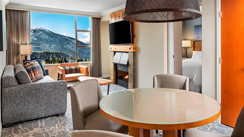 Hotel suite living area with a round glass dining table, sofa, fireplace, and mountain view through a large window.