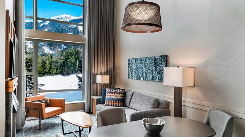 Hotel living area with large windows showing snowy mountains outside; gray sofa, orange chair, round coffee table.