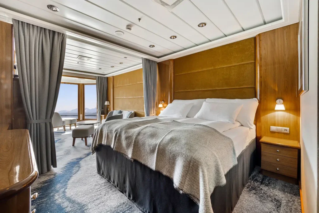 Suite bedroom with a king bed, wood-paneled headboard, grey drapes and a view of the sea.
