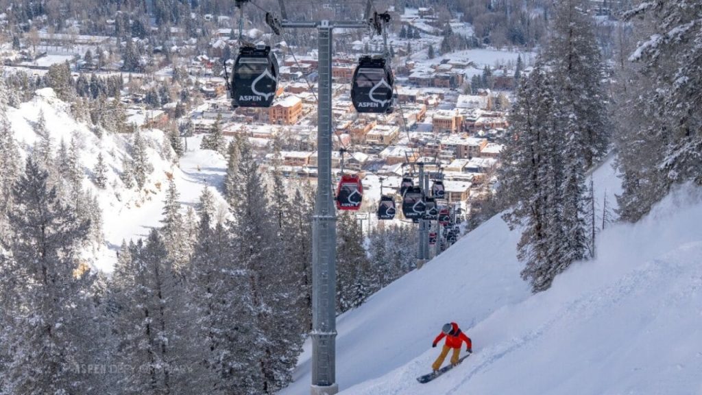 Skier in an orange jacket descending a snowy slope beside gondola cabins above a snowy mountain town.