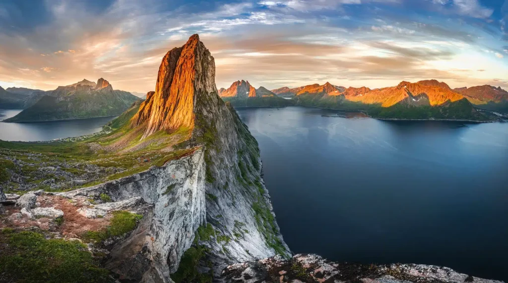 Dramatic rocky peak rising above a fjord at golden hour, with mountains and islands beyond.