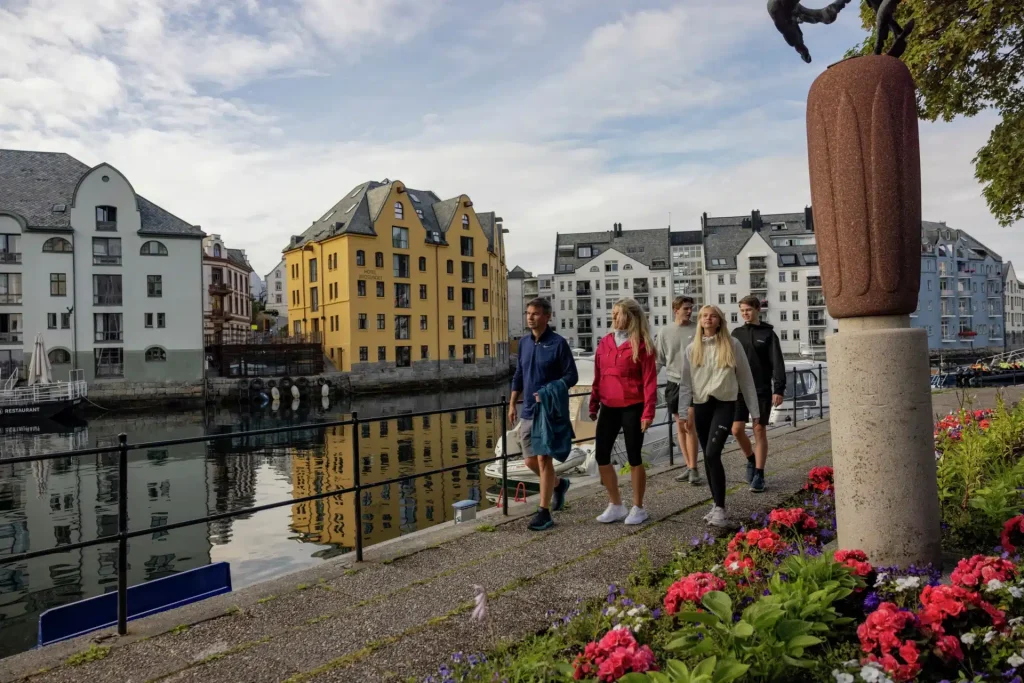 Group of young people walking past colorful waterfront buildings and flower beds in a city.