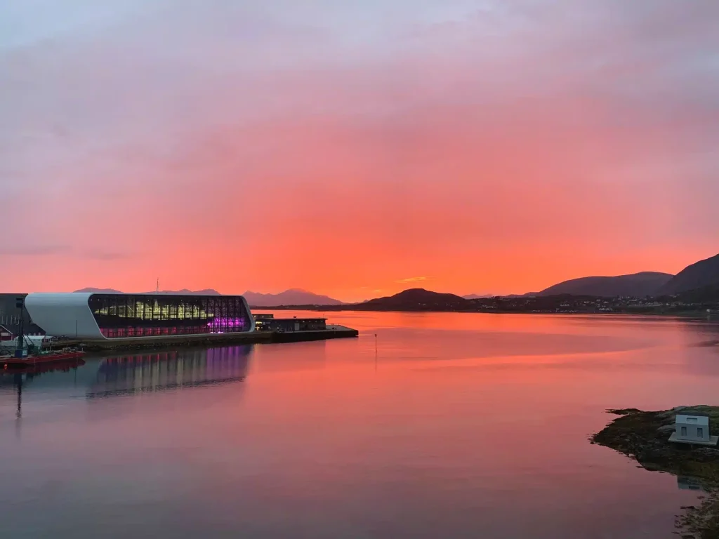 Pink-orange sunset over a fjord with a modern white glass-fronted building on the waterfront.