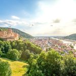 Hilltop view of a red castle ruin above a riverside old town with a stone arch bridge.