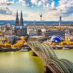 Cologne skyline with twin Gothic cathedral spires, an arched steel bridge over the Rhine, and autumn trees.