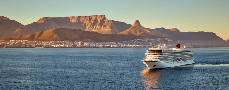 White cruise ship sailing at sunset past a flat-topped mountain range and a coastal city.