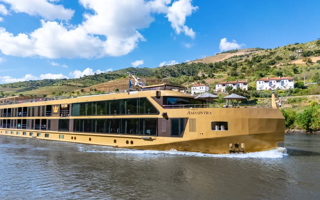 Cruise ship AMASINTRA on a calm river with green hills and a small hillside village in the background under a bright blue sky with clouds.