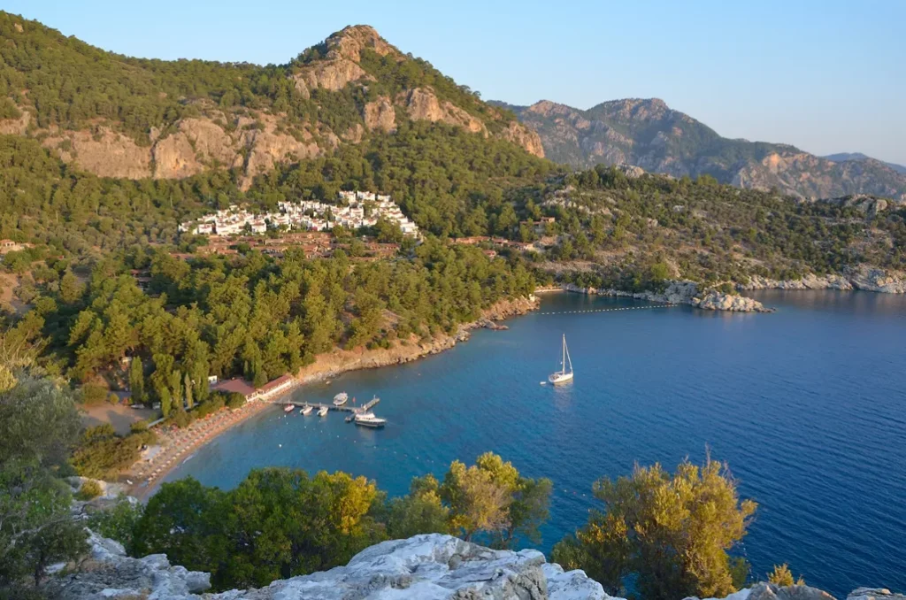 Turquoise bay with a sailboat and a whitewashed hillside village framed by pine-covered mountains.
