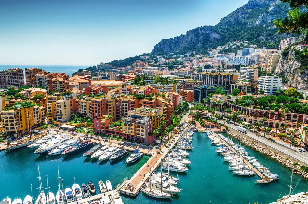 Mediterranean harbor with numerous yachts, colorful buildings, and green hills in the background.