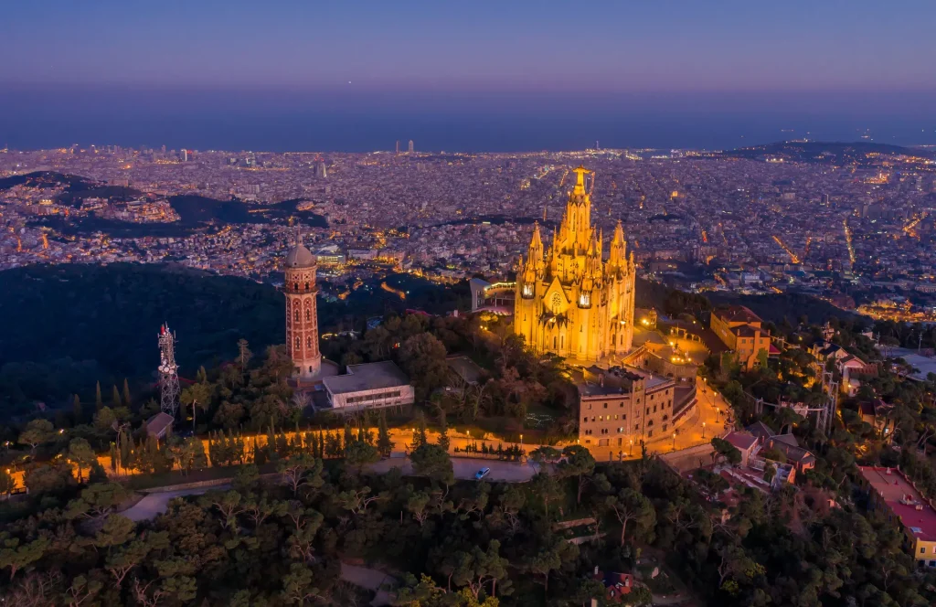 Aerial dusk view of Barcelona's illuminated hilltop church above the sprawling city.