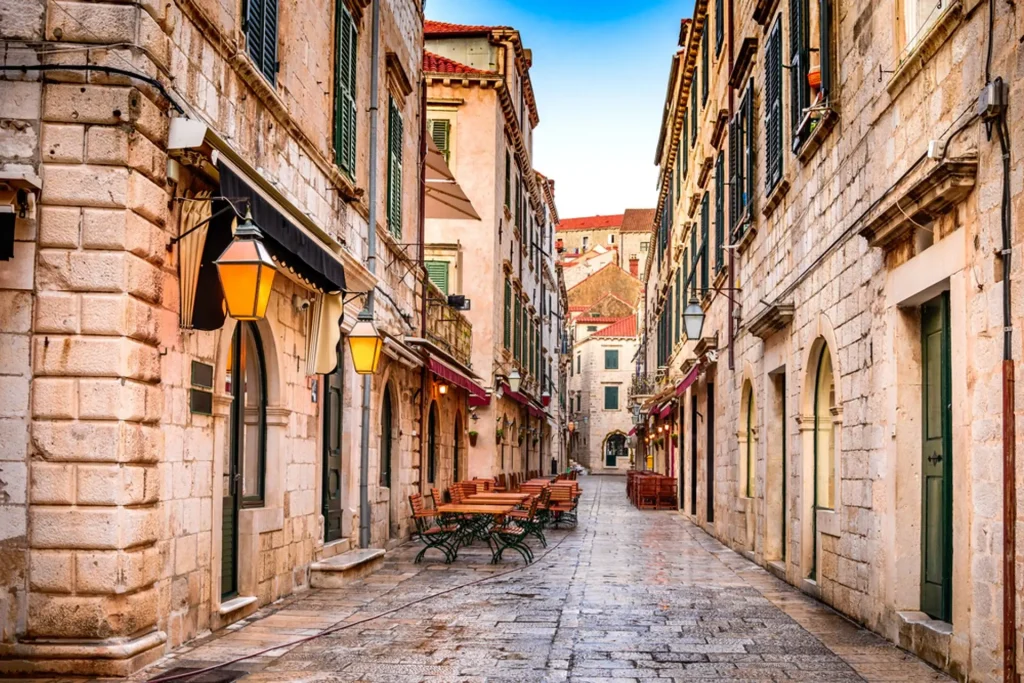 Narrow stone-paved old town street lined with lanterns, cafe tables and shuttered stone buildings.