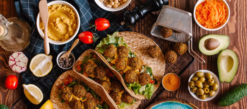 Overhead spread of falafel wraps, hummus, lemons, avocado, olives and vegetables on a wooden table.