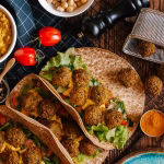 Overhead spread of falafel wraps, hummus, lemons, avocado, olives and vegetables on a wooden table.