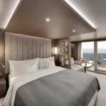 Ship suite bedroom with a large bed, sliding doors to a balcony and an ocean view beyond.