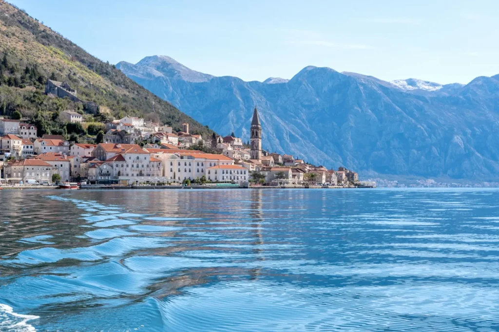 Seaside village with terracotta roofs and a bell tower beneath steep mountains, viewed from the water.