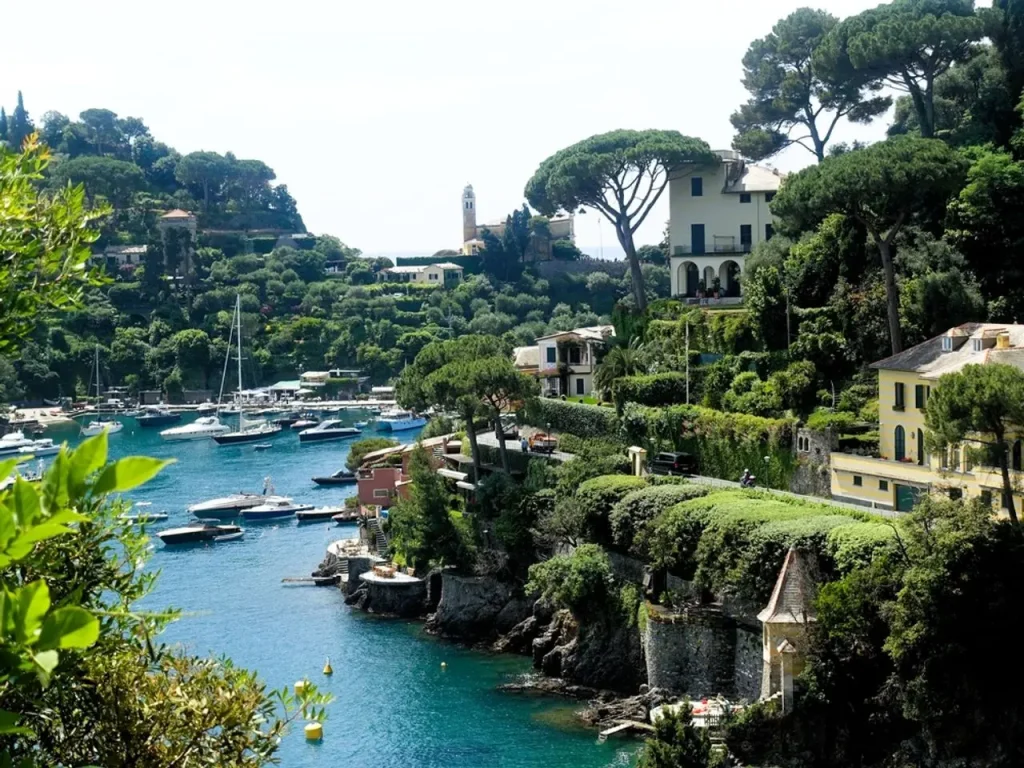 Sheltered bay with moored yachts, turquoise water, and pastel villas and pine trees on the surrounding cliffs.