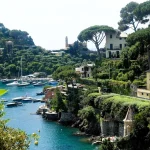 Sheltered bay with moored yachts, turquoise water, and pastel villas and pine trees on the surrounding cliffs.