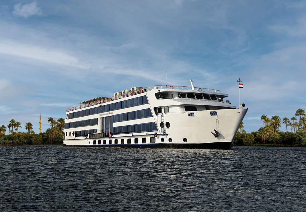 Large white multi-deck cruise ship on dark water, with a palm-lined shoreline and clear blue sky in the background.