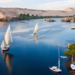 White-sailed feluccas and sailboats on a calm river beside sand dunes and green banks.