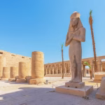 Ancient Egyptian temple courtyard with a tall pharaoh statue, columns, and palm trees.