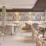 Elegant dining room with cream booths, wood tables, and gold-accent decor; a long mural spans the back wall under soft lighting