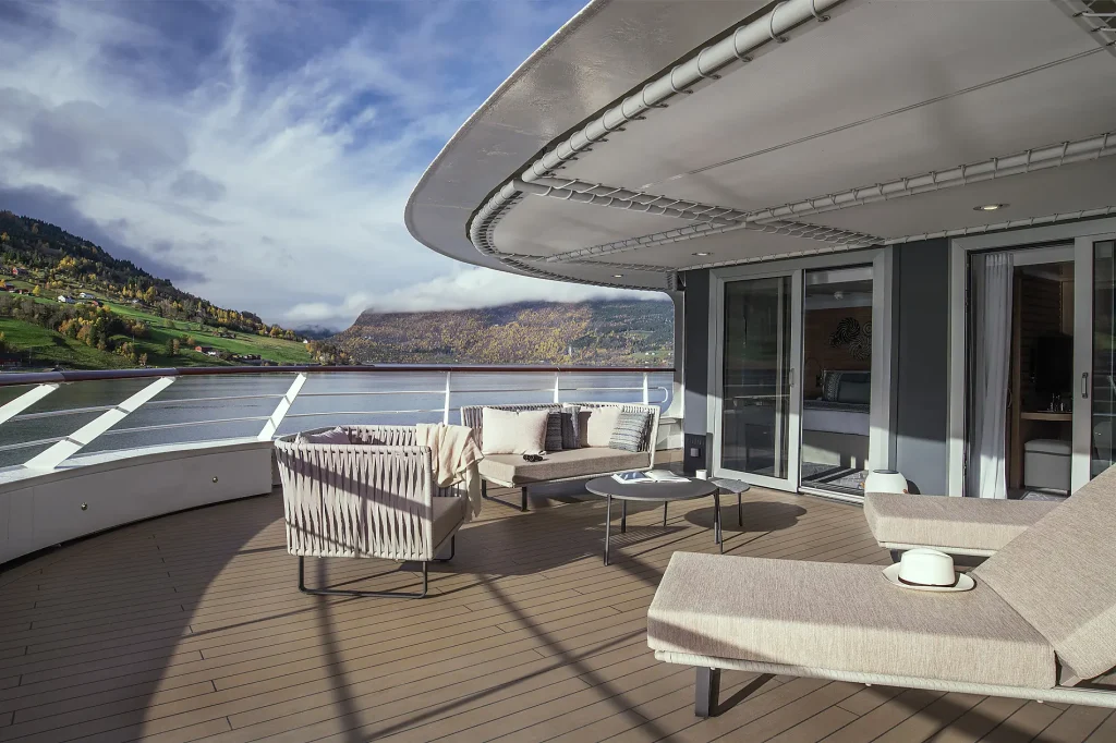 Wide ship balcony with lounge seating overlooking a calm fjord and autumn hillsides.