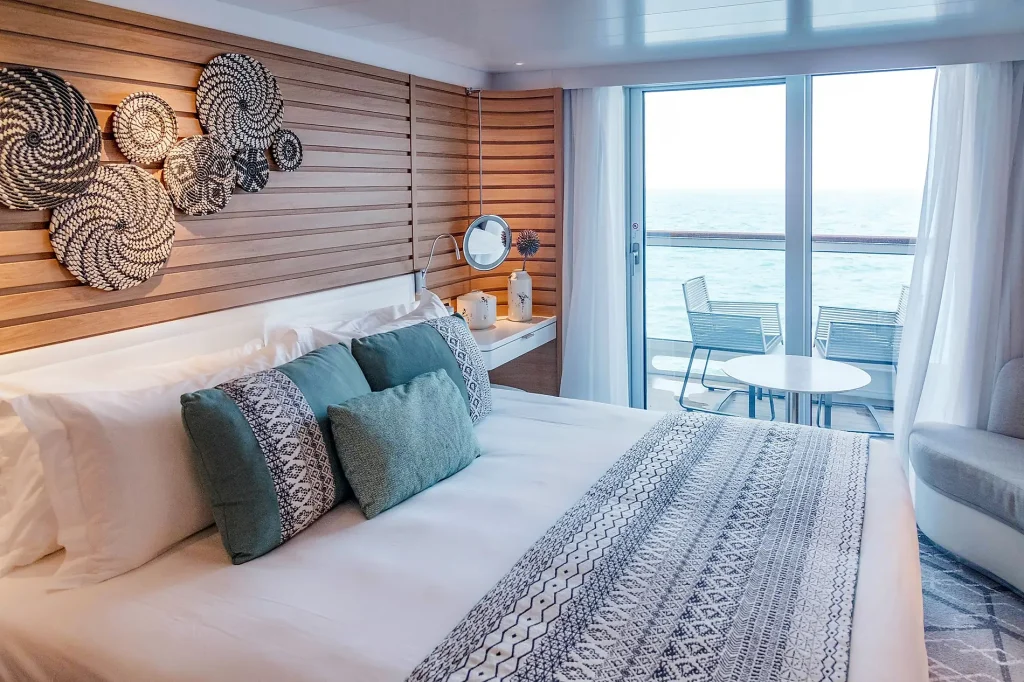 Ship cabin with a bed in patterned linens, teal cushions and balcony doors to the sea.