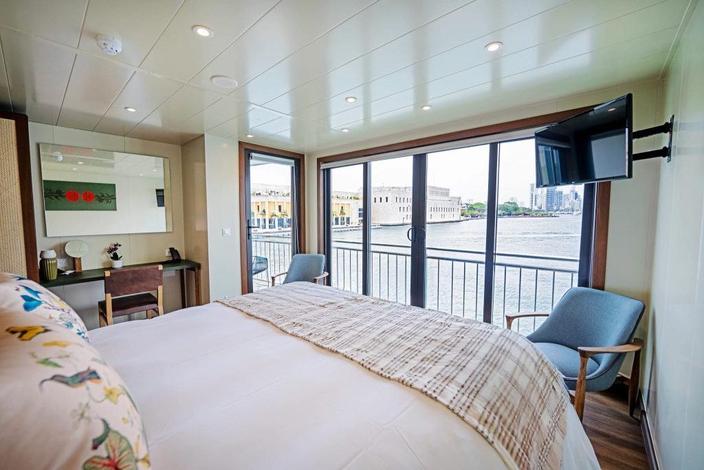 Ship cabin with king bed, balcony doors overlooking a harbor waterfront and a blue armchair.